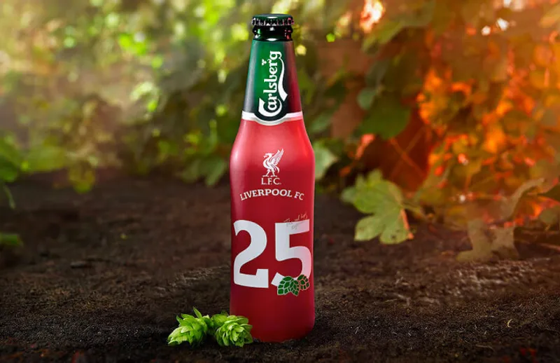 A Carlsberg beer bottle with a special edition red label celebrating Liverpool FC's 25th anniversary. The label features the Liverpool FC logo and the number 25, adorned with hop illustrations. The bottle is set on earthy ground with green foliage in the background, creating a natural and celebratory atmosphere.