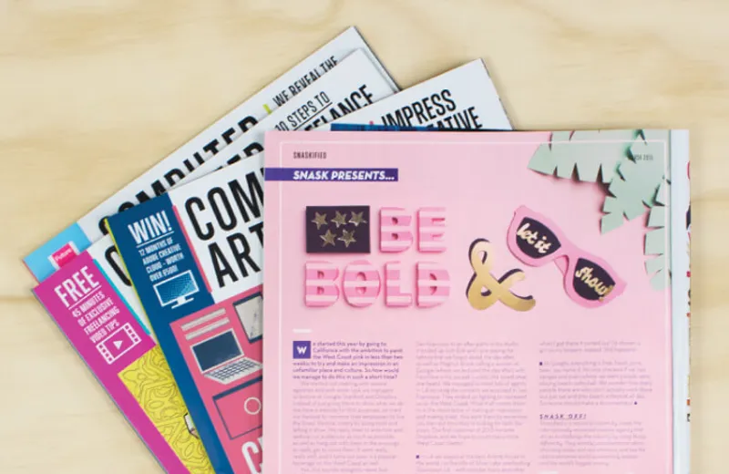 An open magazine spread featuring a pink page with bold text reading 'Be Bold' and an ampersand, alongside pink sunglasses with the words 'let it show.' The page is part of a feature titled 'SNASK Presents.' Several other colorful magazines are partially visible beneath, showcasing creative and artistic themes.