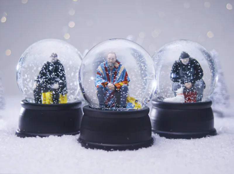 Three snow globes, each containing a miniature figure seated on a yellow bench, surrounded by swirling snowflakes. The central figure wears a colorful jacket, while the others are dressed in darker winter attire. The globes rest on black bases, set against a snowy background with softly blurred lights, creating a whimsical winter scene.