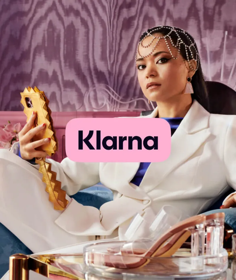 A stylish person in a white suit, adorned with an intricate headpiece, holds a uniquely designed phone case. They are seated in an elegant setting with a purple patterned wall. In the foreground, a glass table displays a pair of transparent-heeled shoes. The Klarna logo is prominently displayed in a pink box, highlighting a blend of fashion and technology.