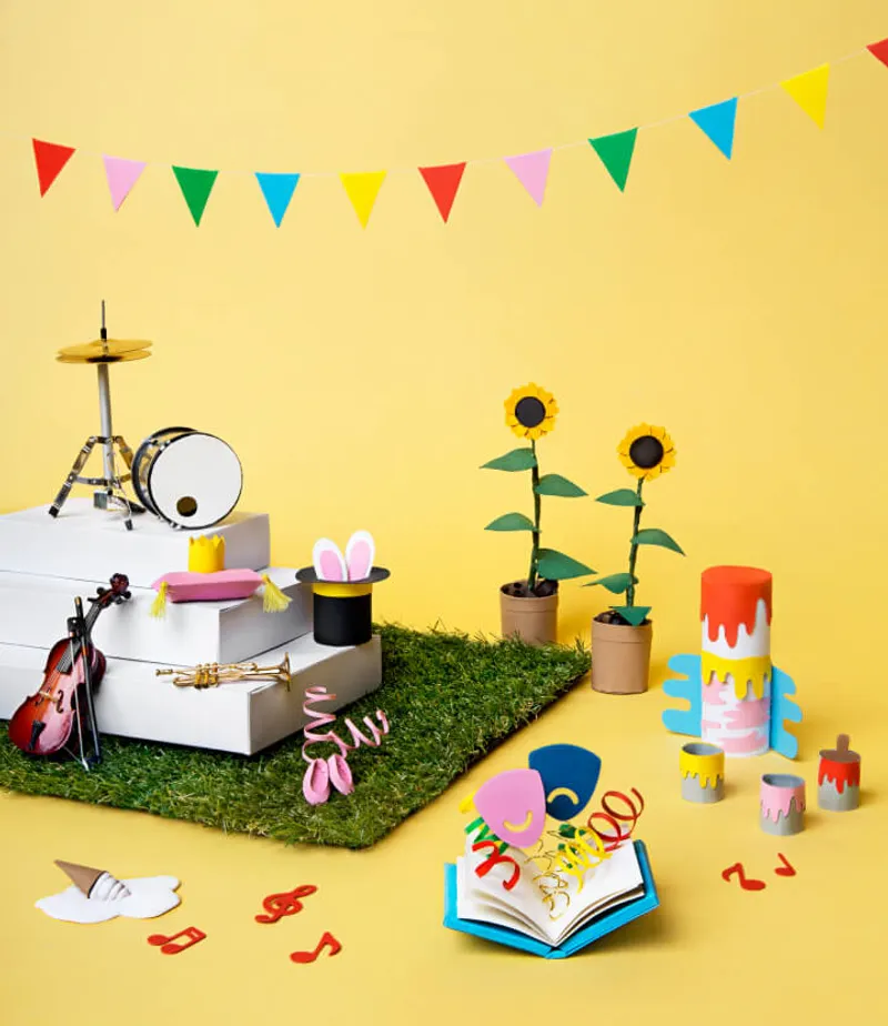 A colorful, playful scene set against a yellow background, featuring a variety of craft items. A drum set and violin rest on white platforms covered with artificial grass. Nearby, a magician's hat with bunny ears, paper sunflowers in pots, and paint cans with dripping colors add to the whimsical display. An open book with paper cutouts and musical notes scattered around complete the festive atmosphere, enhanced by a string of multicolored flags above.