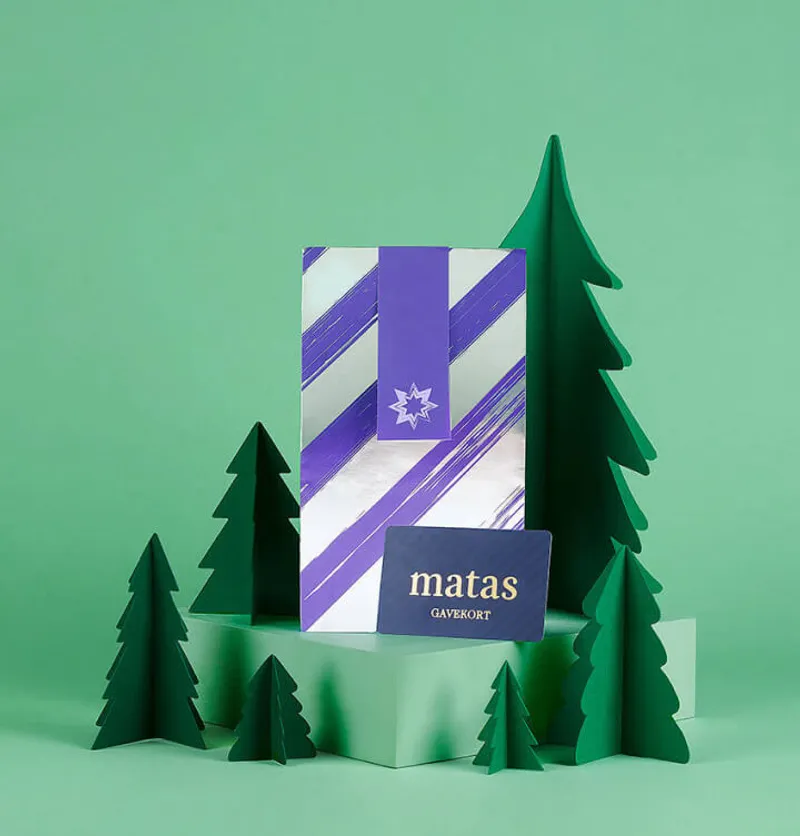 A holiday-themed product photo featuring a Matas gift package in the center, striped in purple and white with a star snowflake emblem. It's displayed on a mint green platform surrounded by stylized dark green paper Christmas trees of different heights. The entire scene has a modern, minimalist design against a matching green background. A navy blue Matas Gavekort label is visible at the bottom of the main package.