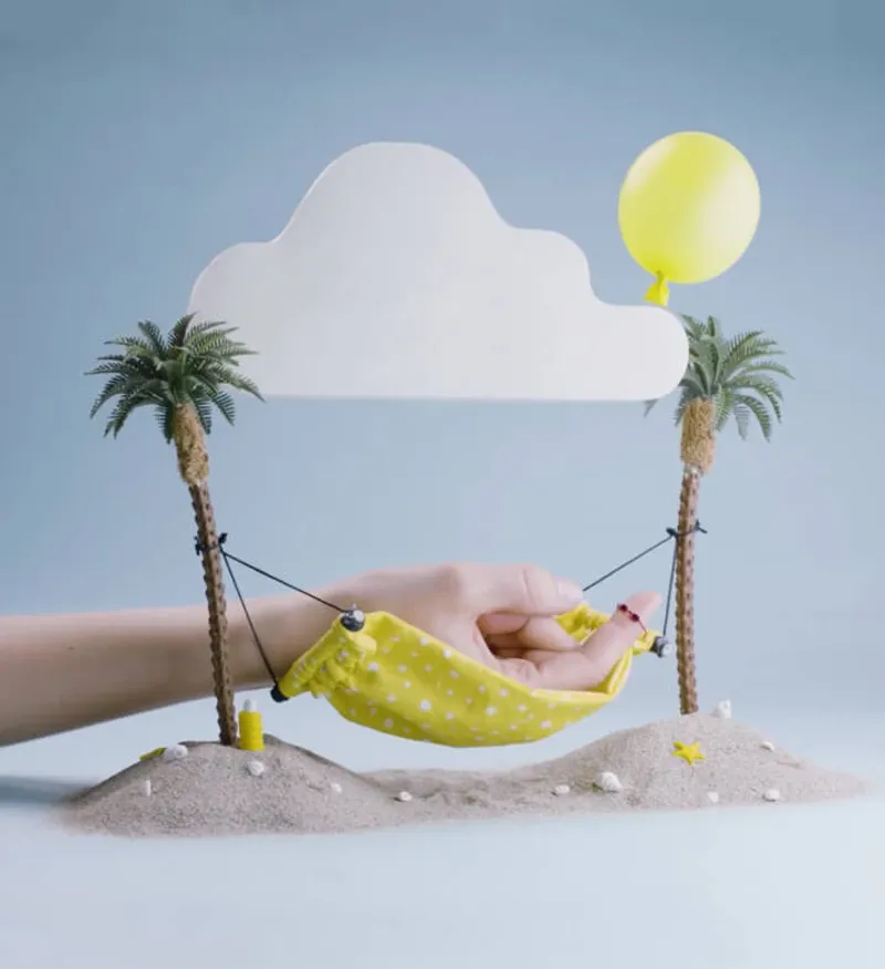 Whimsical design featuring a hand in a yellow polka dot hammock between miniature palm trees on sand, with a cloud cutout and yellow balloon. Showcasing creative and playful design elements.