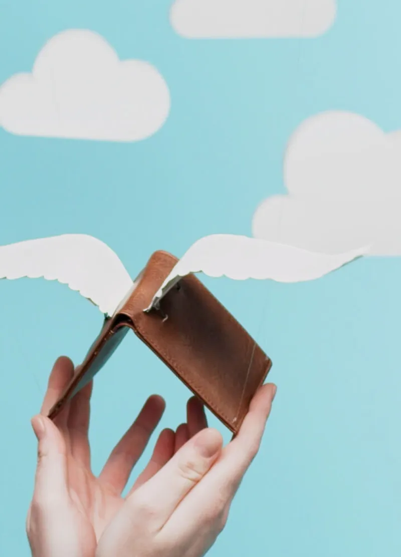 A creative photograph showing hands holding a brown leather billfold with crafted paper wings attached. The background features a soft blue sky dotted with white, fluffy clouds, suggesting effortless movement.