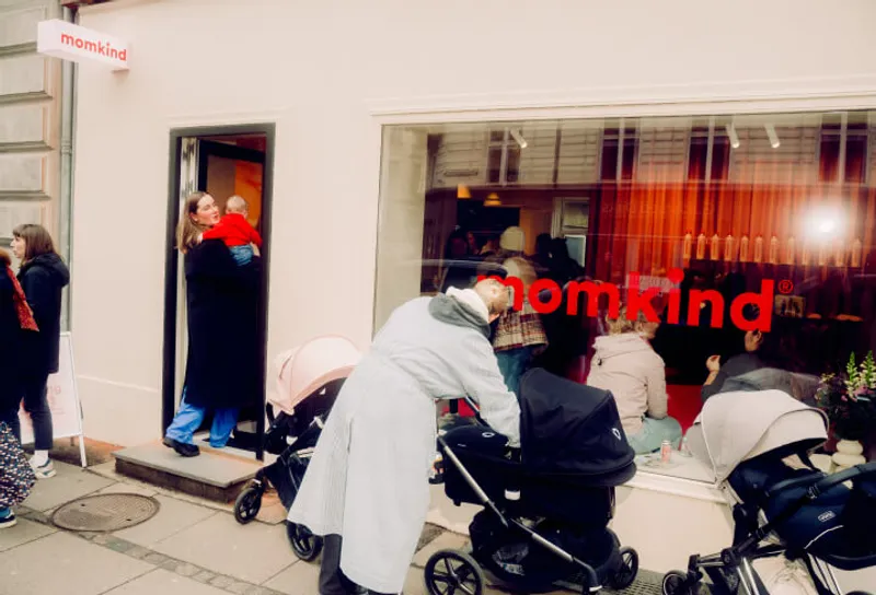 Exterior of a store named Momkind with a group of people and strollers gathered outside. A woman holding a baby stands in the doorway, while others interact near the large window displaying the store's name in red letters.