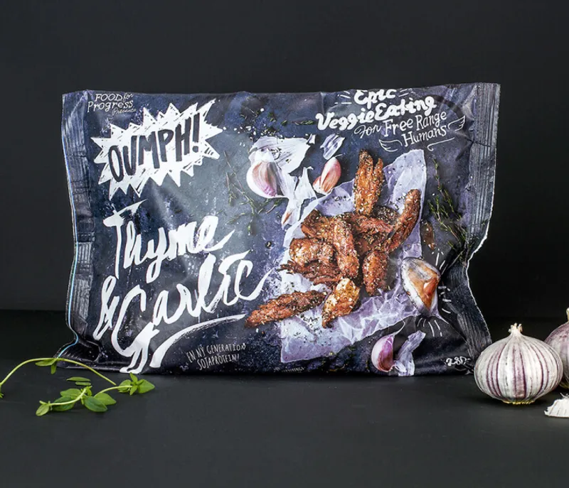 Package of Oumph! Thyme & Garlic, featuring a dark, rustic design with images of seasoned plant-based strips on parchment paper, surrounded by garlic cloves and thyme sprigs. The text highlights 'Epic Veggie Eating for Free Range Humans' and emphasizes a sustainable, flavorful plant-based option. A sprig of thyme and garlic bulbs are placed beside the package on a dark surface.