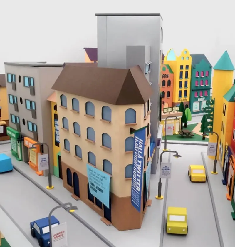 A colorful, miniature model of a city street featuring various buildings with distinct architectural styles. The scene includes a brown-roofed corner building with blue banners, surrounded by other vibrant structures. Small model cars and streetlights line the roads, creating a lively urban environment.