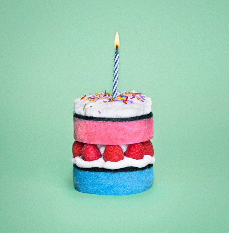 A whimsical cake made from colorful sponge layers, with a blue base and pink top, separated by a layer of raspberries and white frosting. The top is decorated with white icing and rainbow sprinkles, topped with a lit blue and white striped candle, set against a light green background.