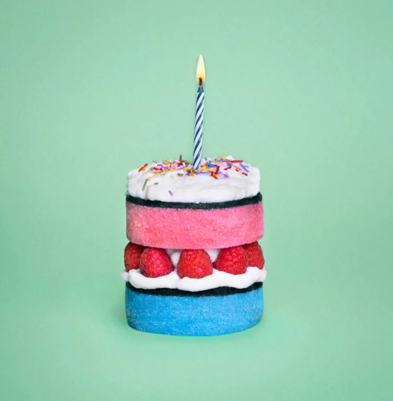 A whimsical cake made from colorful sponge layers, with a blue base and pink top, separated by a layer of raspberries and white frosting. The top is decorated with white icing and rainbow sprinkles, topped with a lit blue and white striped candle, set against a light green background.